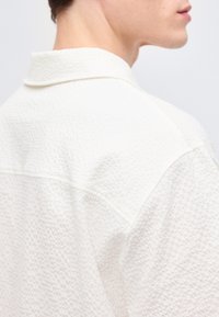 Κοντινό πλάνο στον λαιμό και τον ώμο ενός ατόμου που φοράει υφασμάτινο λευκό ένδυμα με ανεβασμένο γιακά, πάνω σε μονόχρωμο φόντο.