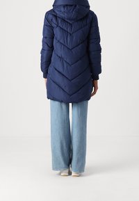 Granatowa kurtka puchowa z kapturem, z wzorem w jodełkę i prążkowanymi mankietami, do tego jasnoniebieskie szerokie spodnie i białe trampki.