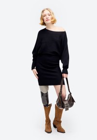 Kobieta o krótkich blond włosach, ubrana w czarną sukienkę z odsłoniętymi ramionami, brązowe buty, trzymająca brązową torebkę, z protezą lewej nogi.