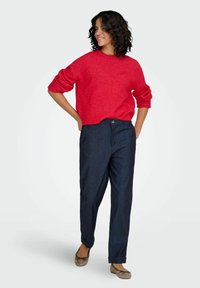 Pull rouge en maille avec manches retroussées, porté avec un pantalon en denim foncé et des ballerines beiges. Coupe décontractée, texture lisse, style casual.