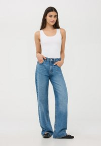 Canottiera bianca abbinata a jeans a gamba larga blu chiaro, con vita alta e classica texture denim. Modella in piedi con le mani nelle tasche.