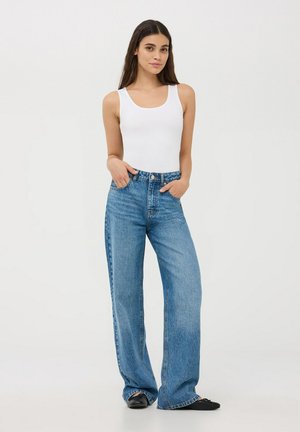Canottiera bianca abbinata a jeans a gamba larga blu chiaro, con vita alta e classica texture denim. Modella in piedi con le mani nelle tasche.