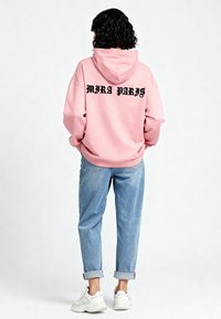 Felpa rosa con "MIRA PARIS" in grassetto nero sulla schiena, abbinata a jeans azzurri arrotolati e sneaker bianche.