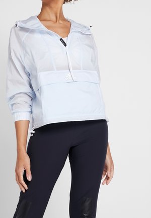 Person iført en lyseblå Adidas quarter-zip vindjakke og sorte leggings mod en ensfarvet lys baggrund.