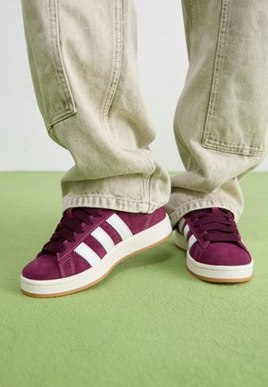 Person iført beige cargobukser og mørkelilla sneakers med hvide striber, stående på en lysegrøn gulv.