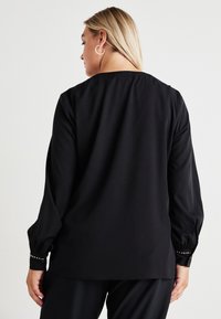 Blusa preta de mangas compridas com decote redondo e mangas bufantes; apresenta detalhes em botão nos punhos; tecido suave com um ligeiro brilho.