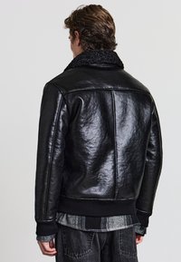 Schwarze Lederjacke mit einer strukturierten Oberfläche, ausgestattet mit einem schwarzen Strickkragen, gerippten Bündchen und Saum. Schlichte Rückseite, keine zusätzlichen Details.