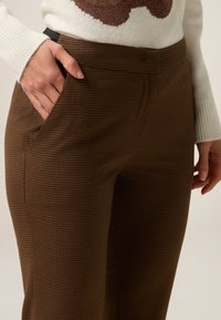 Pantaloni a quadri marroni con una texture liscia, caratterizzati da tasche laterali e un design aderente. Il materiale sembra tessuto e resistente.