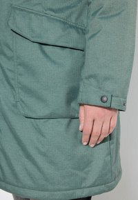Giacca verde, strutturata, con texture, caratterizzata da una tasca laterale e un polsino con bottoni. Il tessuto presenta un leggero motivo a spina di pesce, offrendo un aspetto ordinato.