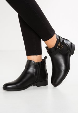 Stivali alla caviglia in pelle nera con superficie liscia, cinturino decorativo e fibbia dorata, caratterizzati da un bordo ondulato e zip laterale per una facile vestibilità.