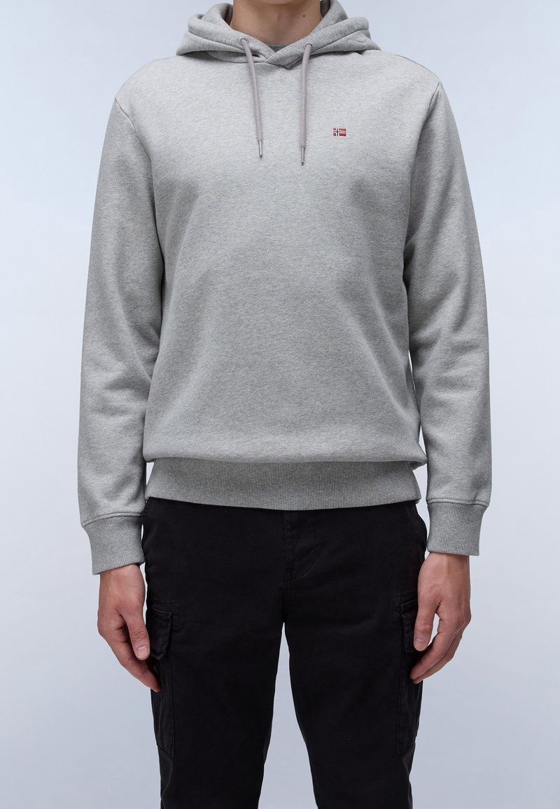 Personne portant un sweat à capuche gris clair uni avec cordons et petit logo rouge, associé à un pantalon cargo noir, debout devant un fond uni.