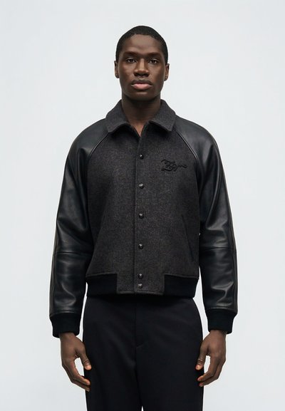 Νεαρός άνδρας φορώντας σκούρο γκρι μπουφάν με κουμπιά και μαύρα δερμάτινα μανίκια, μαζί με μαύρο παντελόνι, στέκεται μπροστά σε απλό φωτεινό φόντο.