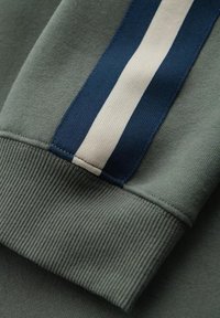 Dunkelgrüner Sweatshirtbund mit strukturiertem Rippenfinish. Beinhaltet ein blaues und cremfarbenes Streifendesign auf einem Akzentpaneel.