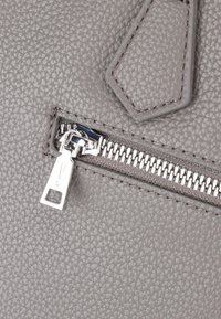 Angolo di una borsa in pelle grigia con texture, dotata di una zip argentata e un tirante, con una superficie decorata.