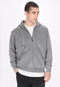 Szara rozpinana bluza z kapturem na sznurki, kieszeniami kangurzymi oraz ściągaczami przy mankietach. Gładki materiał o delikatnej teksturze i luźnym kroju.