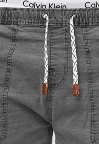 Shorts gris Calvin Klein avec taille élastique à cordon et accents tressés blancs et noirs, ainsi que des détails en cuir marron. Texture en denim lisse.