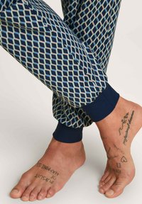 Marineblauwe en lichtblauwe pyjamabroek met een diamantpatroon en elastische boorden. Blote voeten met zichtbare tatoeages op de enkels.