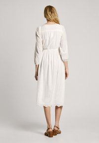 Vit klänning med broderad ögla, trekvartsärmar, elastisk midja och scallopad nederkant, kombinerad med bruna blockklack-sandaler.