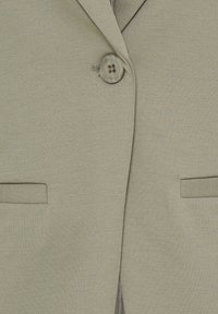 Gros plan sur le devant d'un blazer beige avec un bouton marron fermé et deux poches fendues horizontales de chaque côté.
