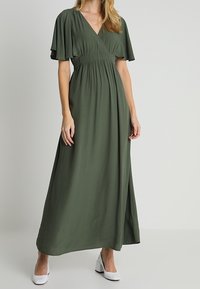 Zelené maxi šaty s překládaným designem, krátkými volánkovými rukávy a elastickým pasem. Hladký materiál; doplněné bílými botami na blokovém podpatku.