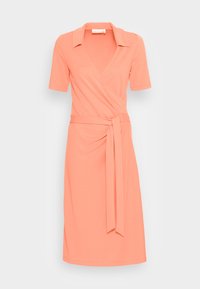 Korálové úzké šaty do délky ke kolenům, s krátkými rukávy, límcem a látkovým páskem uvázaným v pase na jednobarevném světle pozadí.