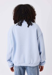 Sweatshirt oversize bleu clair avec ourlet côtelé, texture douce, manches raglan et détail de piping blanc le long des coutures des épaules.