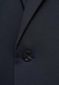 Detaliu al unui blazer din țesătură de culoare bleumarin, cu o suprafață texturată și un nasture negru cu patru găuri cosut pe partea stângă.