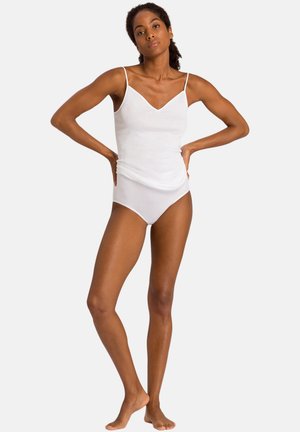 Frau mit dunklem lockigem Haar, die in einem weißen Camisole-Body steht, barfuß mit den Händen in der Hüfte vor einem schlichten hellen Hintergrund.