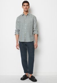 Musterhemd in Weiß mit blauen geometrischen Designs, kombiniert mit dunkelblauen Hosen und schwarzen Slippern. Lässiger Schnitt und hochgekrempelte Ärmel.