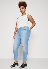 Šviesiai mėlyni siaurėjantys džinsai su pažeistais keliais, derinami su balta be rankovių palaidine ir juodomis atvirų pirštų blokiniais kulnais.