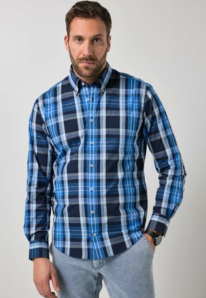 Mand med skæg iført blå og marineblå ternet skjorte med knapper og lyseblå jeans, der står mod en ensfarvet baggrund.