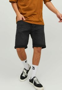 Șorturi negre din denim cu tiv rulat, asociate cu o cămașă maro, șosete albe și adidași negri, evidențiind stilul casual de stradă.
