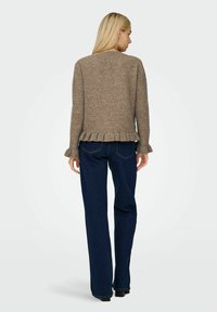 Brauner gestrickter Pullover mit fließendem Saum und Bündchen, kombiniert mit dunkelblauen Jeans, von hinten vor einem hellen Hintergrund betrachtet.