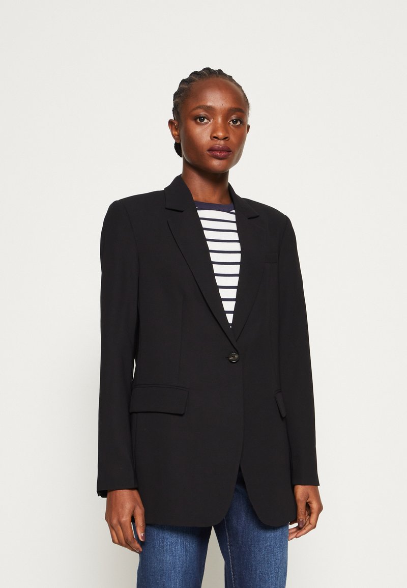 Svart skreddersydd blazer med en enkelt knapp, lange ermer og to frontlommer. Båret over en stripet topp med blå og hvite linjer.
