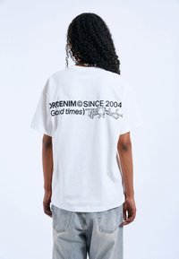 Bílý bavlněný tričko s krátkým rukávem. Na zádech se nachází černý grafický text "ORDENIM SINCE 2004" a "Good times" v různých stylech písma.