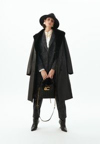 Černý kožený kabát s límcem z umělé kožešiny, vrstvený přes vzorovaný tmavý oblek. Zlaté řetízkové doplňky a texturovaná černá kabelka se zlatým akcentem.