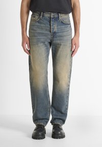 Jeans de mezclilla azul claro con un corte relajado, parte frontal descolorida, diseño estándar de cinco bolsillos y un dobladillo ligeramente deshilachado. Botas negras puestas.