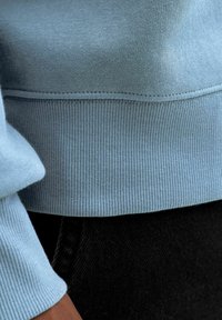 Sudadera azul claro con textura acanalada en los puños y el dobladillo. Tela lisa con un estilo ajustado, combinada con mezclilla oscura.