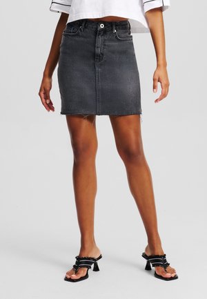 Kvinde iført en falmet sort denim miniskørt med rå kant, kombineret med sorte hælsandaler og en hvid cropped top med sorte striber.