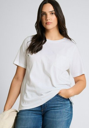 Kobieta z długimi ciemnymi włosami nosi białą koszulkę z krótkim rękawem i niebieskie dżinsy, trzyma beżową kurtkę w lewej ręce, prawa ręka w kieszeni.