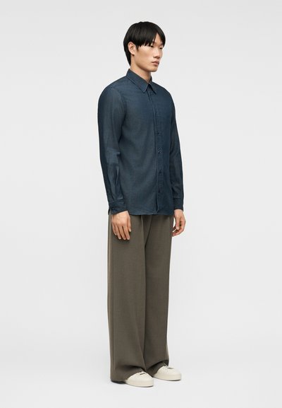 Νεαρός άνδρας που στέκεται στραμμένος προς τα δεξιά, φοράει πουκάμισο σκούρου μπλε με κουμπιά, φαρδιά καφέ παντελόνια και λευκά αθλητικά παπούτσια σε απλό φόντο.