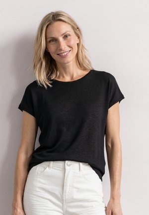 Lyshåret kvinde smilende med skulderlangt hår, iført en sort kortærmet top og hvide bukser, stående mod en ensfarvet lys væg.