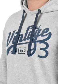 Šedá mikina s tmavě modrými šňůrkami. Obsahuje výrazný grafický text v modré a černé barvě s nápisy "Vintage" a "Established 93." Měkká textura.