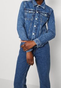 Denim jacket in medium blue with classic collar, button-up front, two chest pockets, and long sleeves, featuring visible stitching details.