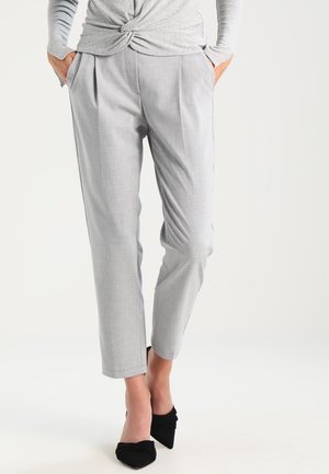 Frau trägt hellgraue, maßgeschneiderte, knöchellange Hose mit Händen in den Taschen und schwarze spitz zulaufende Schuhe vor weißem Hintergrund.