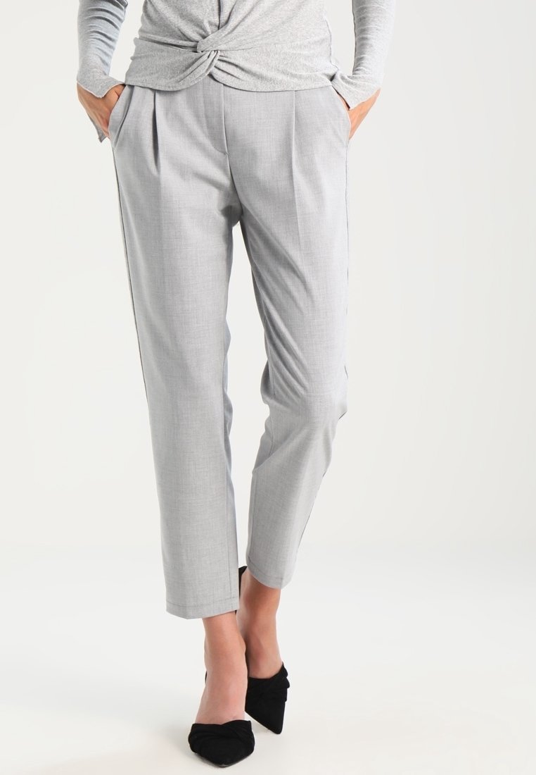 Kobieta w jasno szarych, dopasowanych spodniach długości do kostek, z rękami w kieszeniach, nosząca czarne szpilki z ostrym noskiem na białym tle.