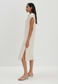 Langes, beiges, ärmelloses Kleid mit lockerer Passform, seitlichem Schlitz, glatter Textur und minimalistischer Gestaltung. Kombiniert mit flachen Sandalen.