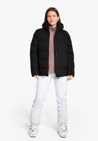 Schwarze Steppjacke mit Kapuze, mit einem gesteppten Design und Reißverschluss, getragen über einem rosa Fleece-Oberteil und weißen Hosen.