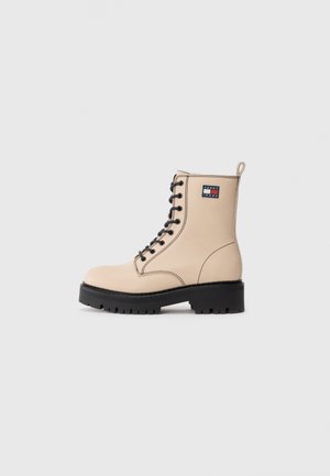 Beżowy skórzany botek za kostkę na sznurowadła z czarną podeszwą i naszywką z logo Tommy Jeans po bokach, prezentowany na jasnoszarym tle.