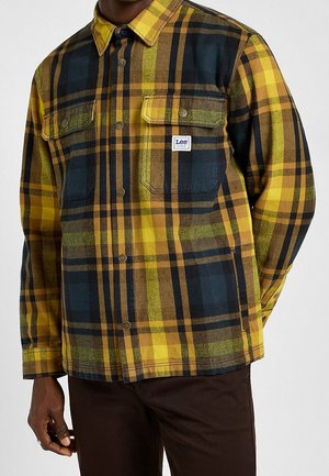 Homme portant une chemise à carreaux jaune, verte et bleu foncé à boutons, avec deux poches poitrine et une étiquette de la marque Lee sur la poche gauche.